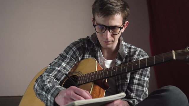 young man writing song lyrics with acoustic guitar in cozy room. close-up.