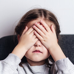 Little girl covers face with hand. Sad offended child girl sitting at sofa at home. Child abuse.