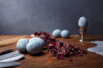 Blue painted easter eggs, one egg in egg cup, dry Hibiscus tea and silver decorative feathers on wooden background, copy space