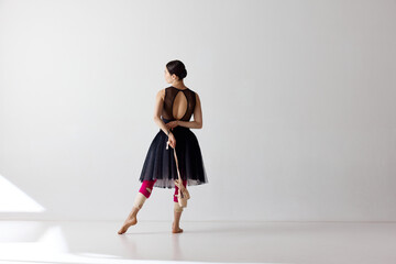 Back view. Young and graceful ballet dancer wearing black dress posing and holding pointe shoes on hand over white background © Lustre Art Group 