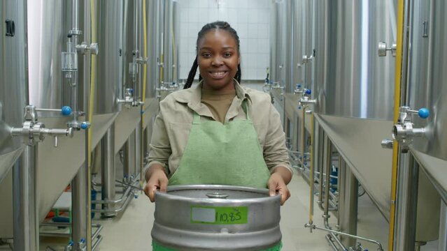 Medium portrait shot of African American female brewery worker in apron standing between beer fermentation tanks, lifting up empty keg from floor, looking at camera and smiling