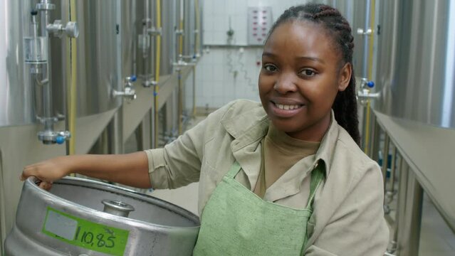 Chest-up portrait shot of happy African American female brewery employee in apron, with braided hair standing next to large fermenter tanks, holding empty beer keg, looking at camera and smiling