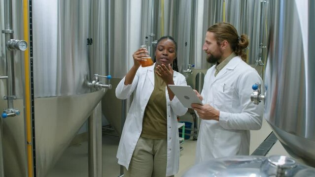 Medium full shot of two multinational brewery technicians in white coats standing in fermentation and storage facility, holding flask with beer sample, and discussing its characteristics and colour