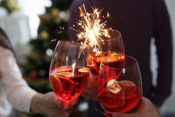 People hold glasses with cocktails with sparklers in front of Christmas tree closeup