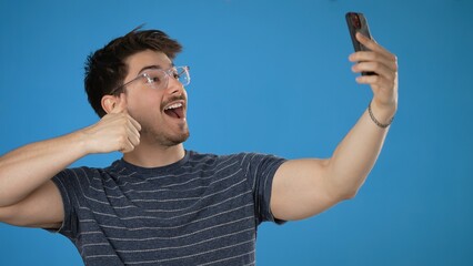 Fototapeta premium Fun young brunet man 20s wears striped t-shirt purple shirt doing selfie shot on mobile cell phone post photo on social network meet greet wave hand isolated on blue background studio portrait