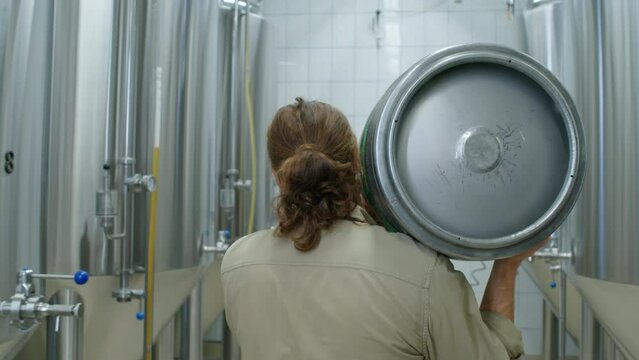 Medium rear follow shot of brewery worker with curly red hair carefully carrying heavy full beer keg on shoulder on production floor between rows of tall stainless steel fermentation tanks