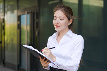 Successful beautiful business woman standing against backdrop buildings writes notes in a notebook or plans a working day in business clothes.