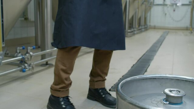 Medium shot of two full beer kegs on floor in brewery, male worker walking up, picking one and carrying it away, and large steel fermenter tanks in background
