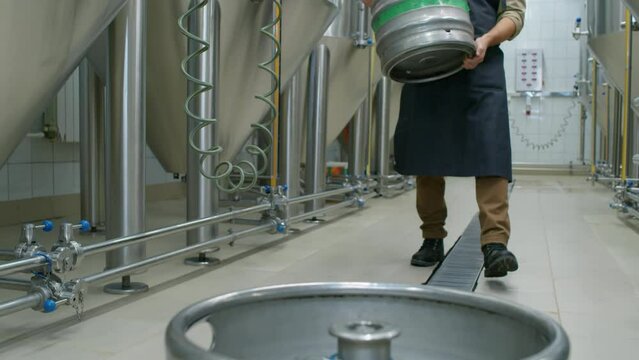 Full shot of young Caucasian man at work in craft brewery, walking along rows of fermentation tanks, carrying empty beer keg, and setting it down on floor next to another one