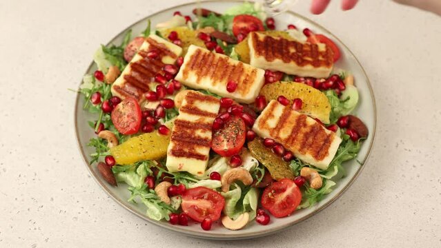 Warm salad with fried halloumi cheese, nuts (cashews and almonds), cherry tomatoes, pomegranate, orange and arugula. Traditional Mediterranean cheese. Woman pouring olive oil on salad.