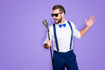 Portrait of handsome attractive singer in blue pants with suspenders and black glasses, singing hit with open mouth in microphone gesture with hand isolated on grey background