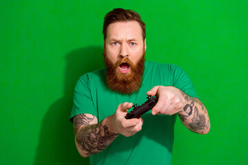 Portrait of masculine man red beard wear trendy t-shirt holding playstation joystick play excited game isolated on green color background - obrazy, fototapety, plakaty