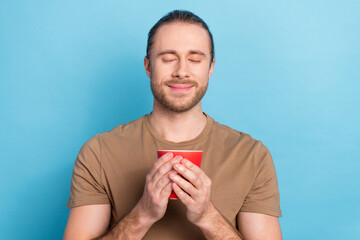 Photo of cheerful relaxed handsome man tail hairdo dressed beige t-shirt hold mug enjoy morning time isolated on blue color background