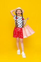 A little girl holds shopping bags. A child smiling in a summer hat and shorts is engaged in children's shopping. A happy child in full growth on a yellow isolated background is holding onto a hat.