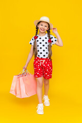 A little girl holds shopping bags. A child smiling in a summer hat and shorts is engaged in children's shopping. A happy child in full growth on a yellow isolated background is holding onto a hat.