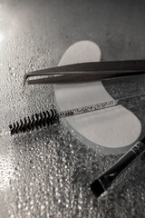Black cosmetic brush for eyebrows and eyelashes, eyebrow tweezers and patches lie on a wet table