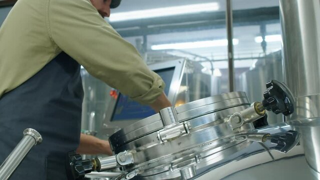 Medium tilt shot of hands of Caucasian male brewery worker unscrewing knobs on sight glass, opening hatch and looking inside large steel tank at beer wort brewing process