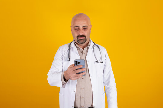 Using Smartphone, Close Up Portrait Of Caucasian Doctor Using Smartphone. Isolated Yellow Background, Copy Space. Middle Aged Bald Hospital Professional Medical Physician Holding Phone With One Hand.