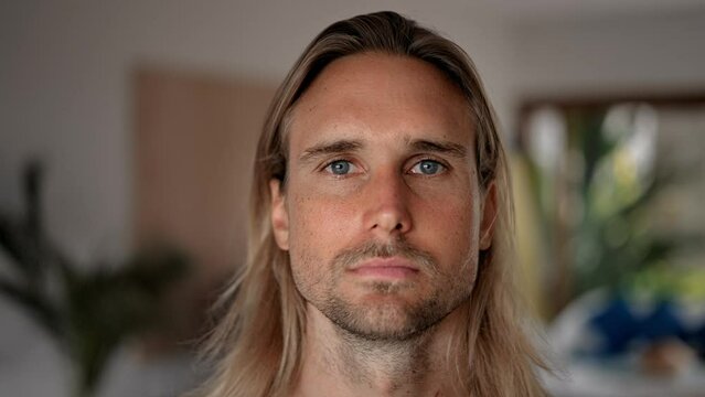 Unhappy male person face home portrait. Blue eyes looking at camera close up. Sad bristle surfer man long hair. Upset shy guy. Kind casual people. Young adult hippie inside house. Hipster hairstyle.