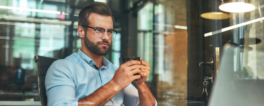 Fresh coffee. Portrait of young and successful bearded man holding cup of coffee and looking at laptop while sitting at his working place