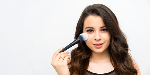 Young woman applies powder on soft facial skin with cosmetic brush. Brunette lady does professional makeup standing on grey background with copy space