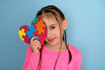 A teenage girl holds an autism symbol in her hand, closing one eye with a postcard and looking into the camera with a sweet smile. Hand made card of colored puzzles