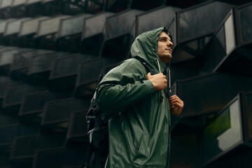 A man in a green rain jacket standing in front of a futuristic dark building in heavy rain wearing...