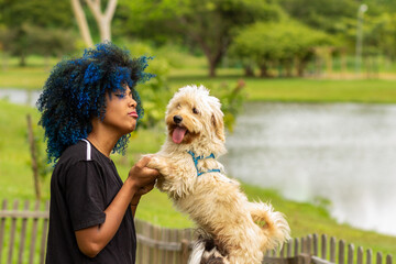 Uma jovem negra de cabelos tingidos de azul, segurando as patas dianteiras de seu cachorro que está parado, em um parque bem arborizado, desfocado ao fundo.