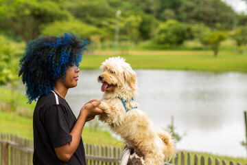 Uma jovem negra de cabelos tingidos de azul, segurando as patas dianteiras de seu cachorro que está parado, em um parque bem arborizado, desfocado ao fundo.