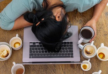 Exhausted female worker surrounded by coffee cups sleeping at workplace over laptop. Tired over...