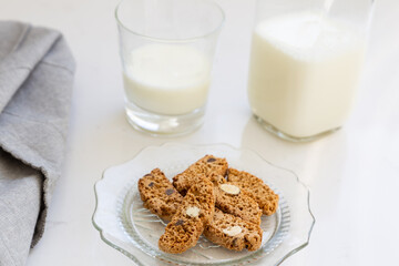 Selective focus high angle view of almond and chocolate biscotti in vintage scalloped glass plate, with grey serviette and bottle and small glass of milk in soft focus background