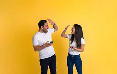 Cheerful couple dressed in white t-shirts screaming and giving high five to each other while using smart phones isolated on yellow background
