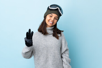 Mixed race skier girl with snowboarding glasses over isolated blue background happy and counting three with fingers.