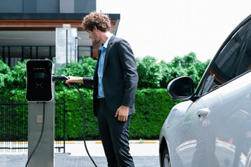 Progressive businessman with electric car recharging at public charging station at modern city...