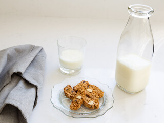 Selective focus view of almond and chocolate biscotti in vintage scalloped glass plate, with grey serviette, bottle and small glass of milk in soft focus background