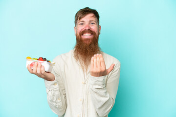 Redhead man with long beard holding a bowl of fruit isolated on blue background inviting to come with hand. Happy that you came