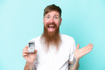 Redhead man with long beard holding a engagement ring isolated on blue background with shocked...