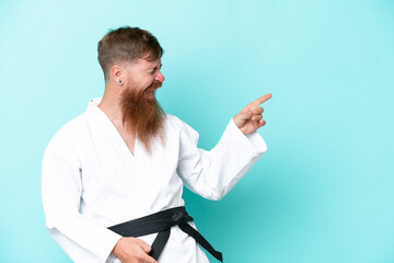 Redhead man with long beard doing karate isolated on blue background pointing finger to the side and presenting a product