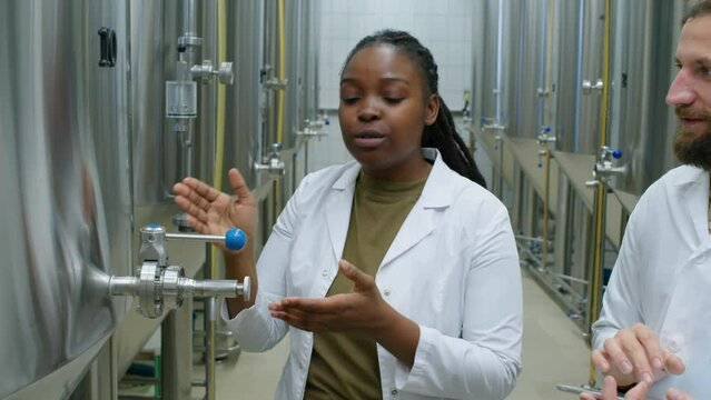 Waist-up shot of African American female craft brewery technologist in white coat teaching Caucasian male trainee about configuration and functions of beer brewing equipment