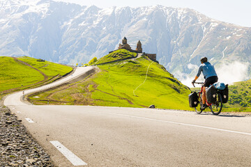 Male cyclist on touring bicycle cycling towards Gergeti trinity church with mountains background....