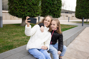 Charming smiling little girls taking selfies with a smartphone while sitting on a bench in the park. Technologies of modern communication.