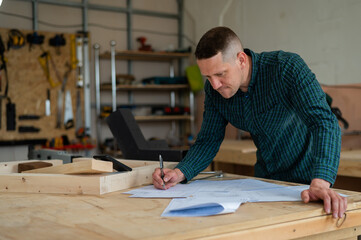 Portrait of a carpenter in a plaid shirt draws a workshop blueprint. 