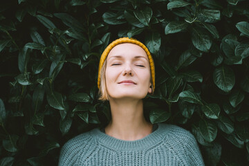 Portrait of relaxed and confident woman with eyes closed on background of green leaves wall. Thoughtful person in front of green hedge. Joy, zen and balance people. Stability through mental health - obrazy, fototapety, plakaty