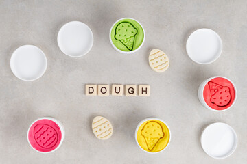 Obraz na płótnie Canvas Colorful dough in white jars on gray background. Word DOUGH written with tile letters. Flat lay