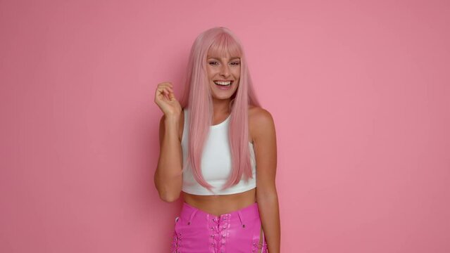 Pretty girl with pink hair standing in the midle of pink background, raises one hand to the ear and smiles happily showing satisfaction, having a good time, copy space, high quality video