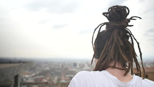 A look from behind a young guy with dreadlocks and beads around his neck, dressed in a white T-shirt, looks thoughtfully at the city from a high balcony in the open air. Slow motion, beautiful people