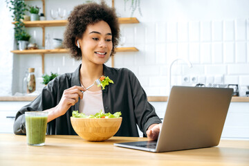 Lovely positive african american young woman, sitting at table in kitchen, eating fresh vegetable salad, watching healthy food or home workout videos on laptop, smiling. Healthy lifestyle concept