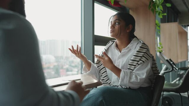 Confident Indian woman discussing something with her colleague near window in the office