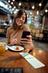 Young woman at coffee shop, looking at phone and smiling. Cheerful happy girl enjoying her time.