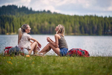 Young female friends are sitting on the ground beside the lake while hiking the hills. Trip, nature, hiking